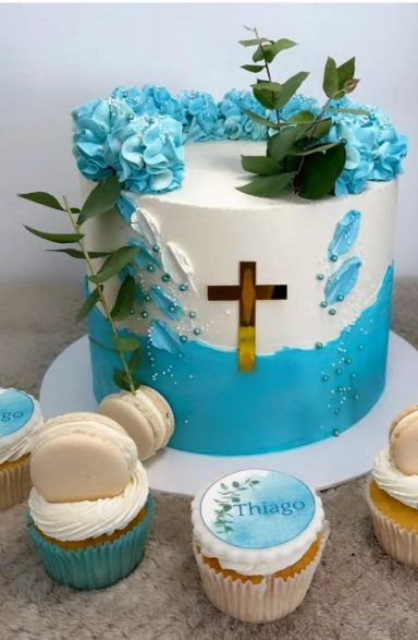 TARTA DE COMUNIÓN Tarta blanca decorada con flores azules, hojas y un crucifijo, acompañada de cupcakes.
