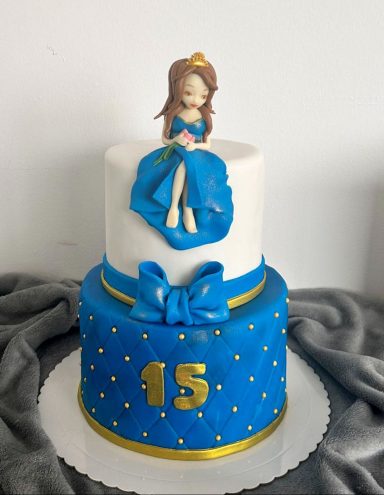 TARTA PARA 15 AÑOS Tarta de dos pisos azul, con figura de una princesa y decoraciones doradas.